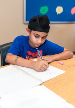 A middle eastern boy in a blue t-shirt diligently writes in a classroom, focusing on his work.