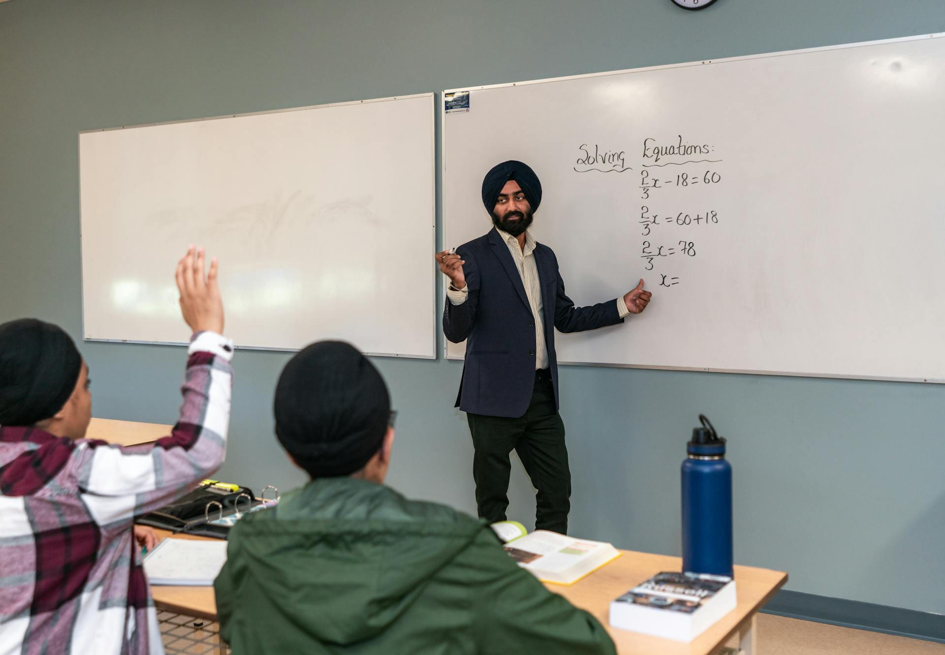 Middle Eastern male teacher in classroom instructing students on solving mathematical equations on whiteboard.