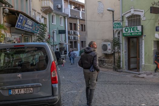 A man walks on a cobblestone street in Izmir, Turkey, surrounded by buildings and a parked Peugeot car.