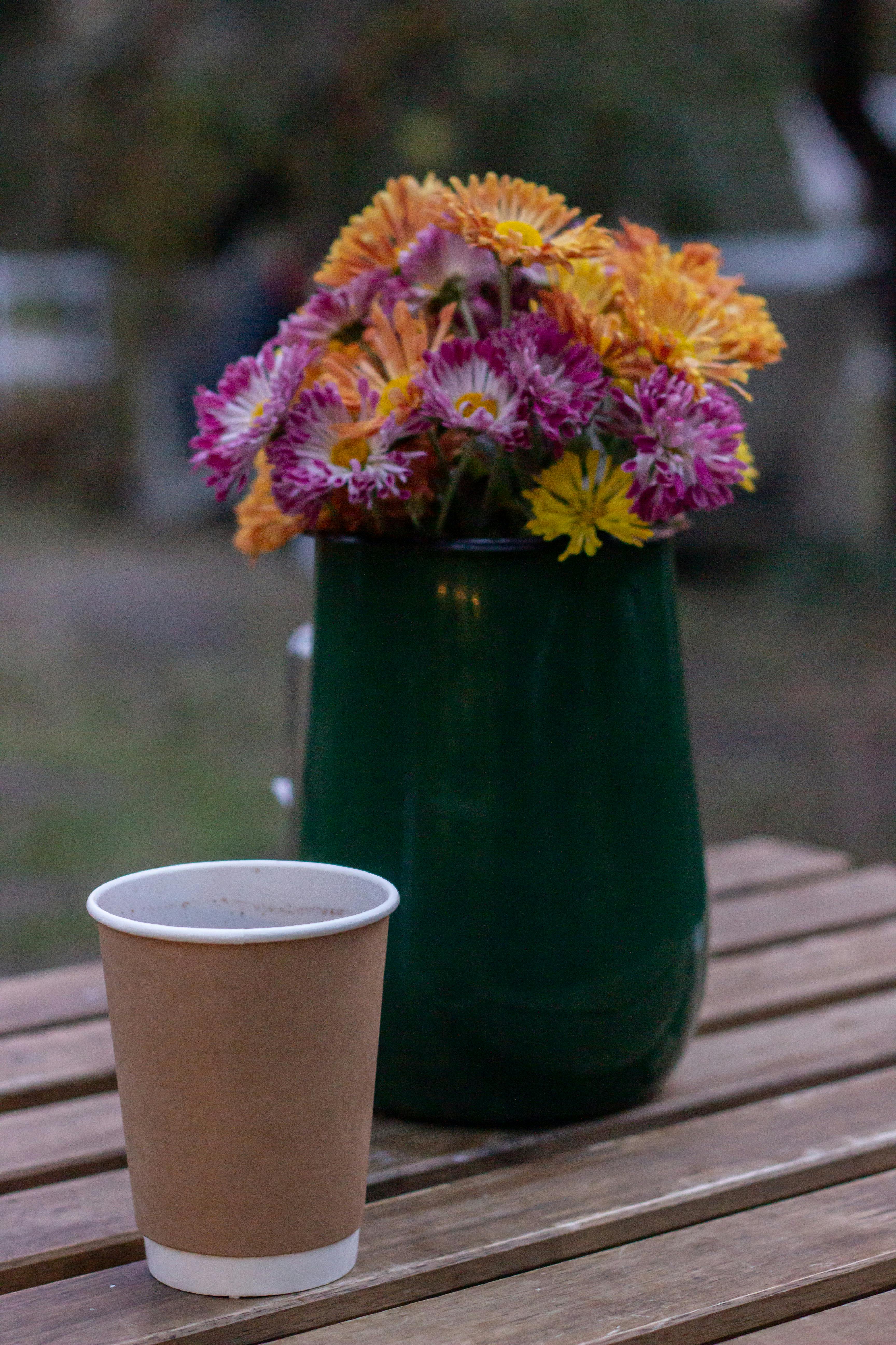 Colorful fall flowers in a green vase next to a coffee cup on an outdoor wooden table.