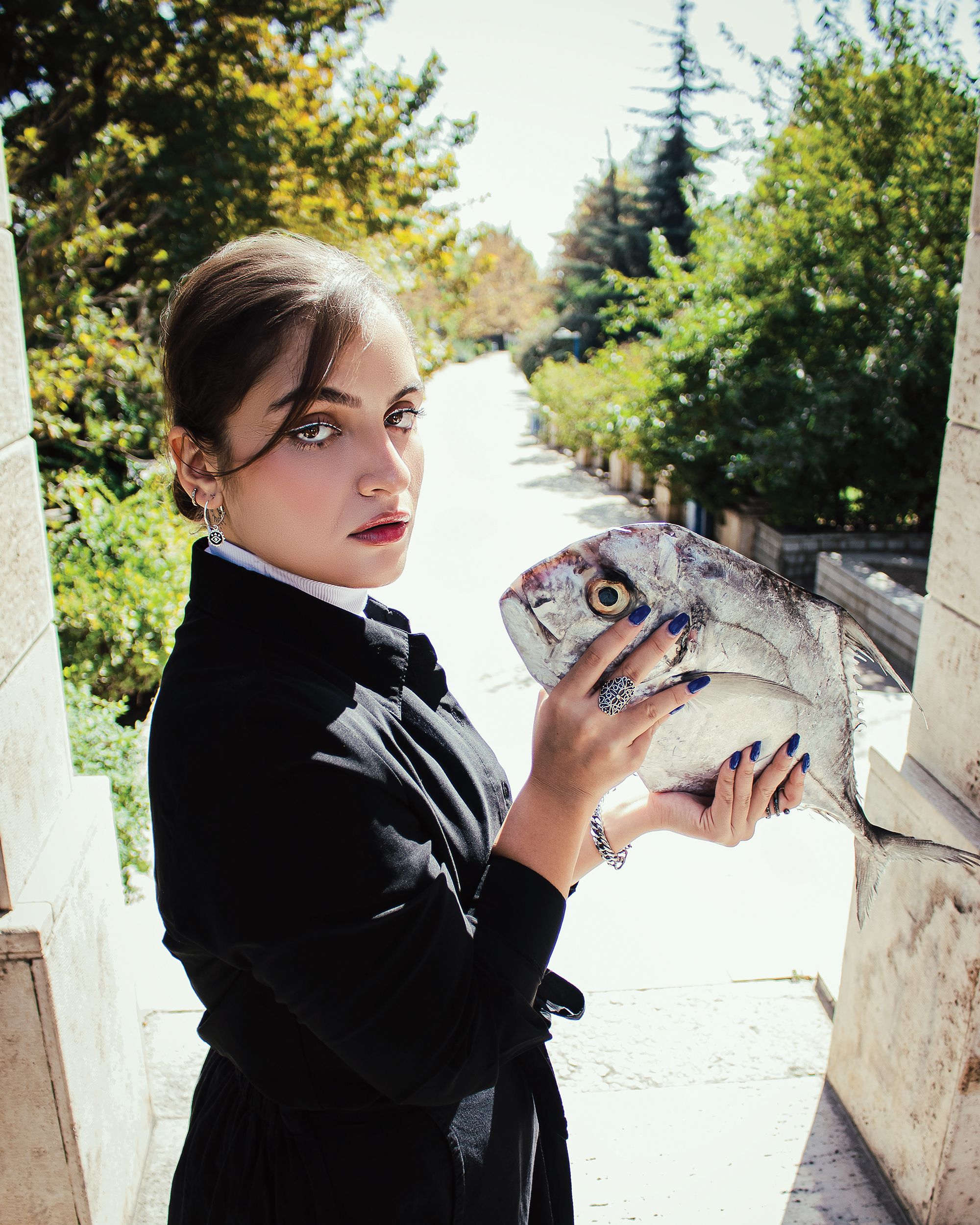 A stylish woman in a black coat holding a fish, posing outdoors in Tehran's summer setting.