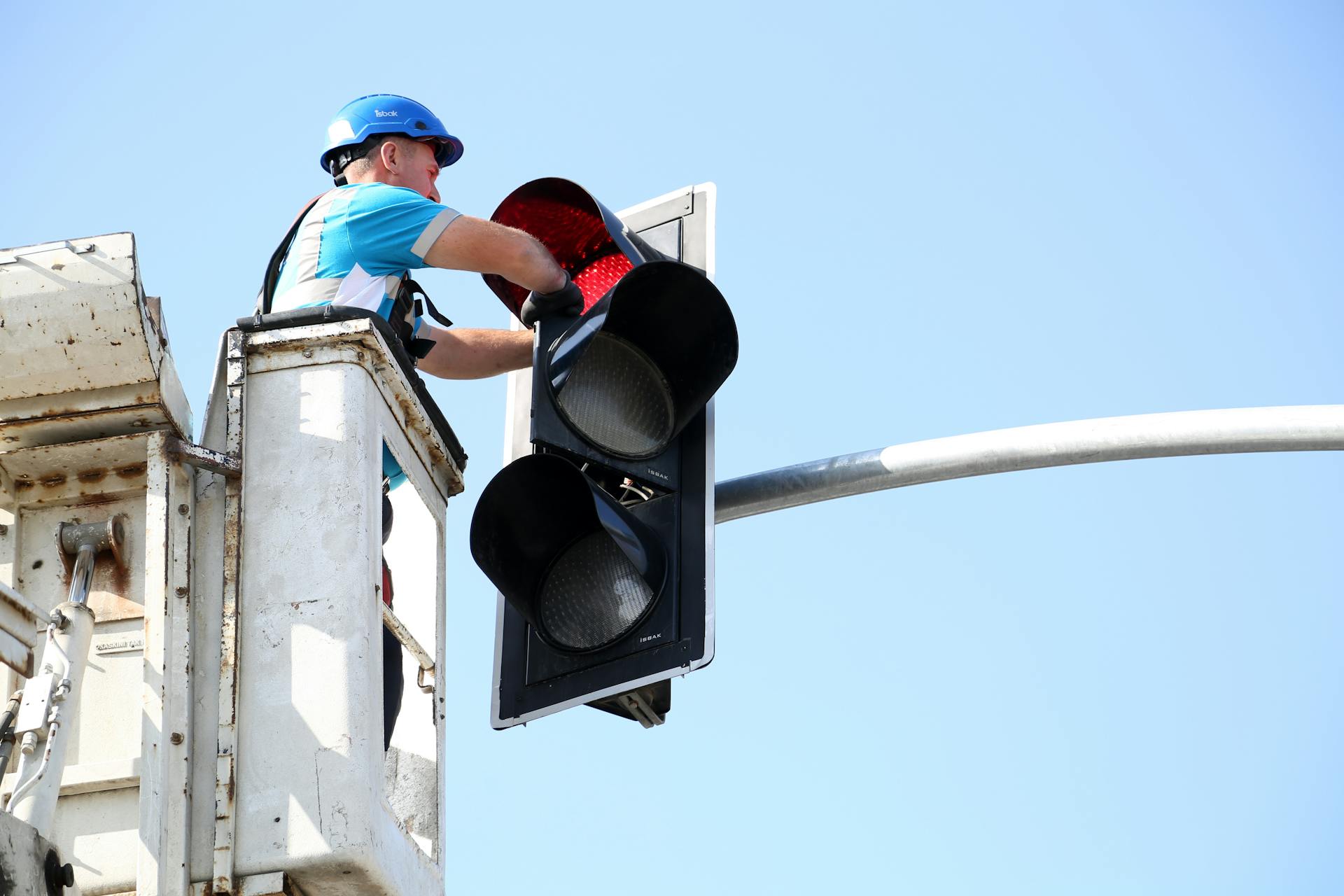 A worker in a helmet fixes a traffic light from an elevated platform on a sunny day.