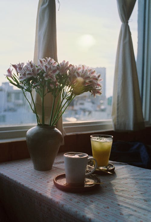 Tea, Coffee and a Vase with Blooming Flowers on a Table Standing by a Window