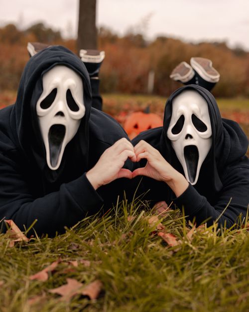 Free Couple lying on grass in Ghostface masks, creating heart shape with hands. Halloween fun in the fall season. Stock Photo