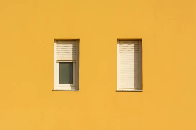 Deux fenêtres d’une façade jaune, le volet roulant de gauche arrêté à mi-course et celui de droite entièrement descendu