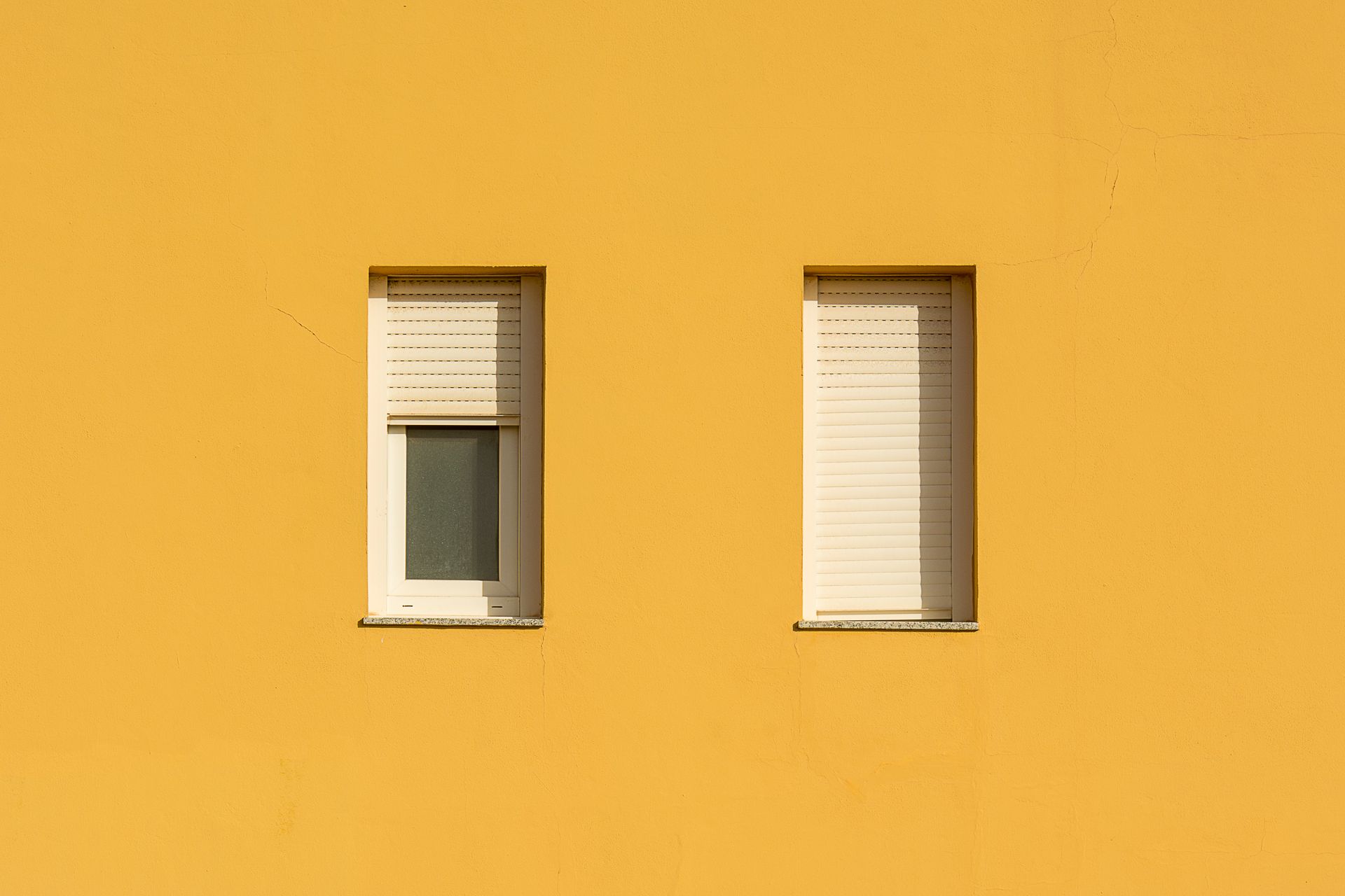 A yellow wall featuring two closed windows with roller shutters, showcasing a minimalist design.