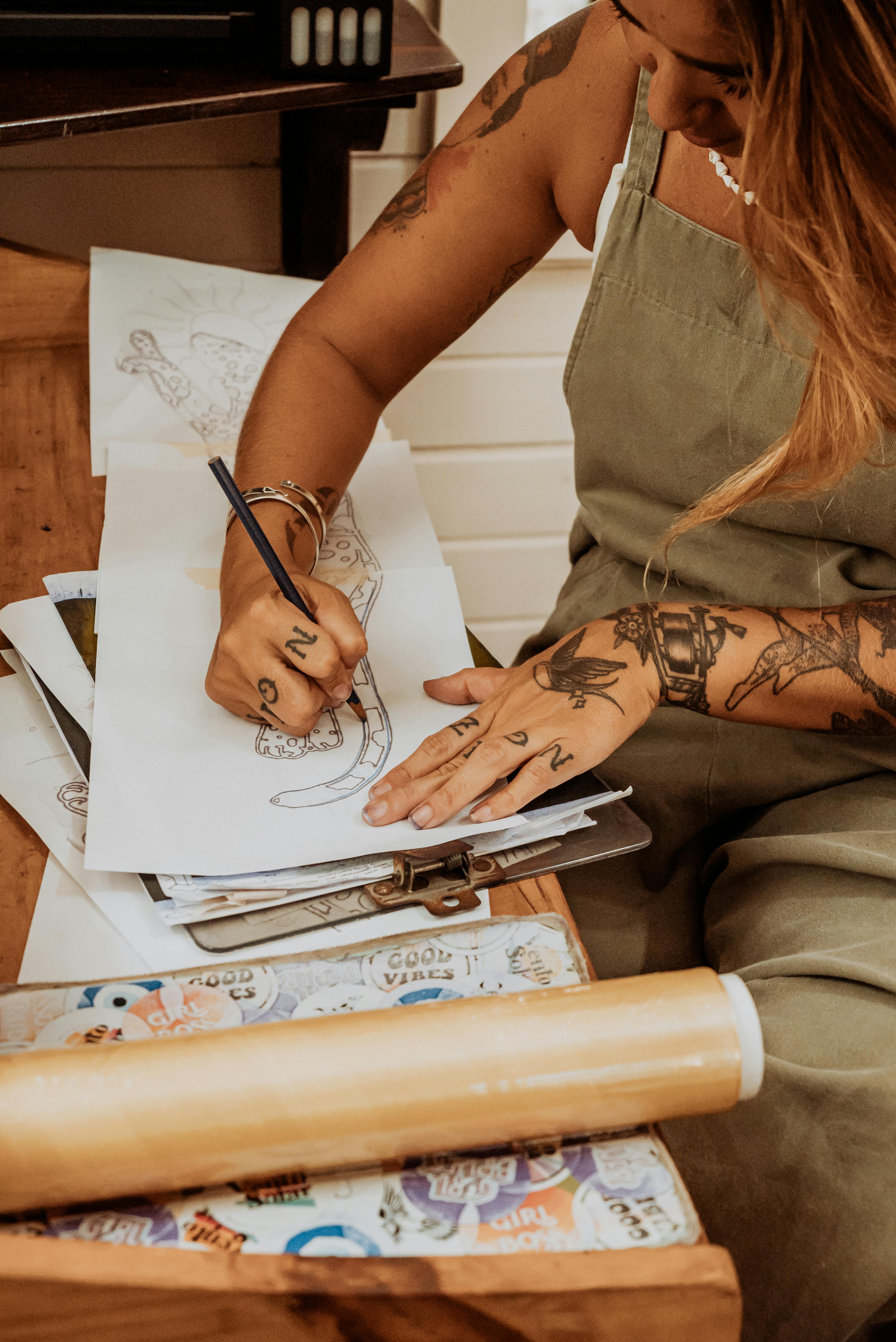 A tattooed woman artist sketching detailed designs in a cozy studio setting.