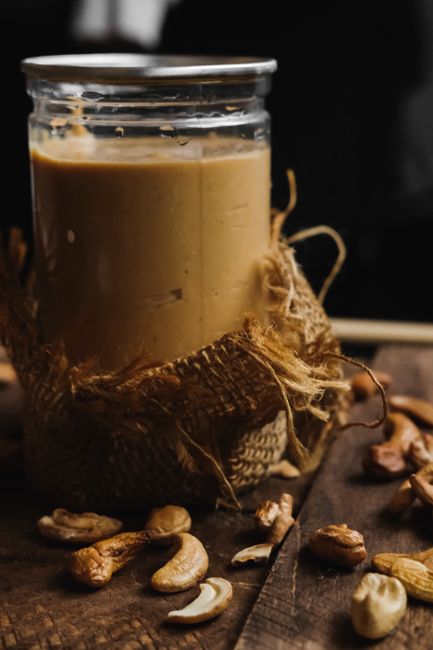A glass jar of pale, creamy cashew butter with a spoon lifting a swirl out of it, surrounded by whole raw cashews on a wooden board.