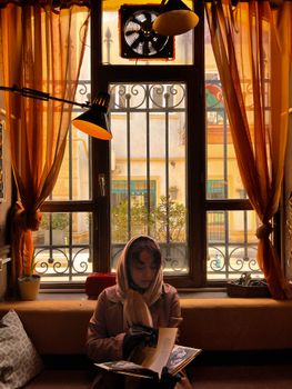 A woman in a hijab reads quietly by a sunlit window, creating a warm, serene atmosphere.. woman, hijab, reads