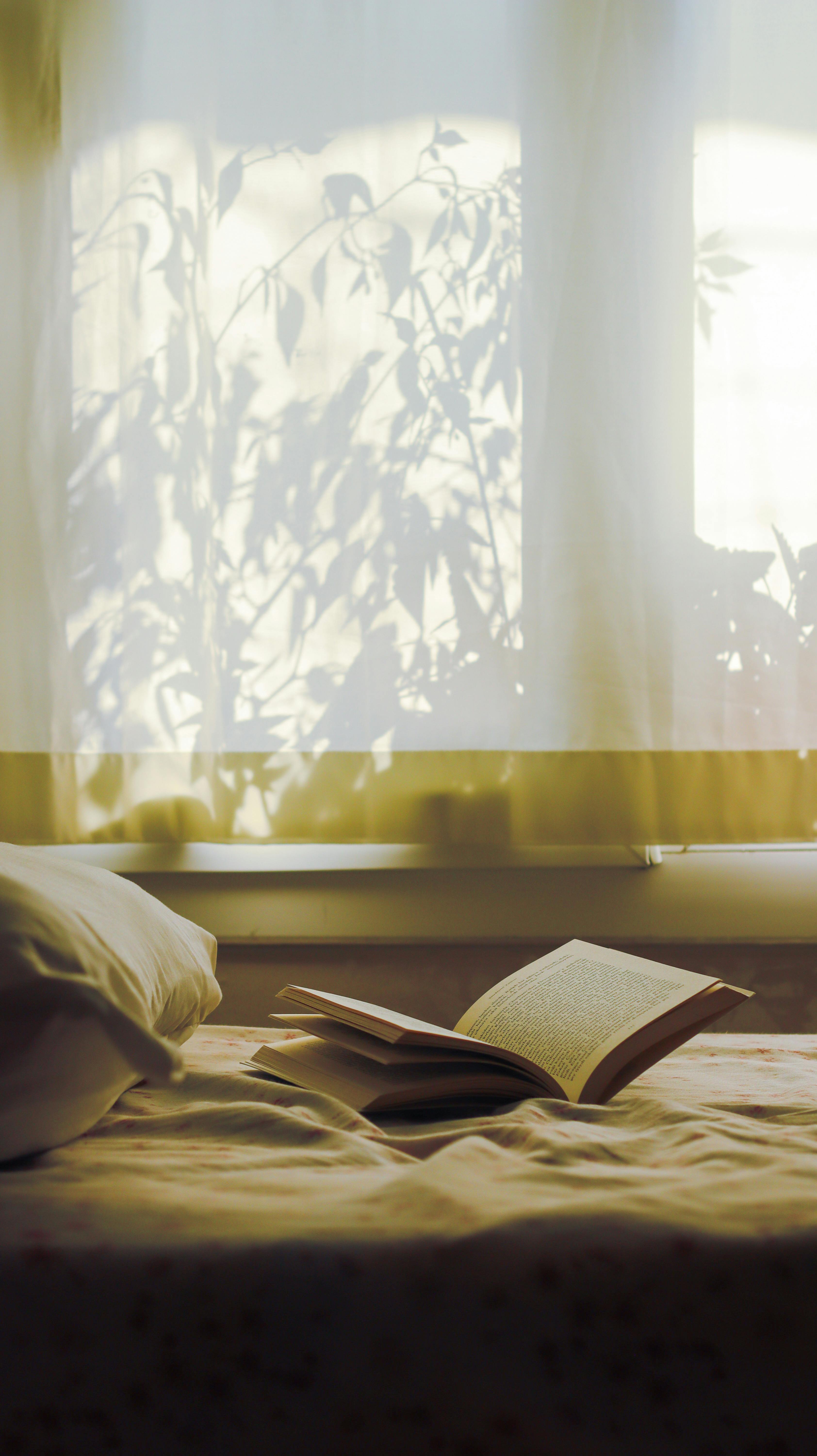 An open book lies on a bed with sunlit curtains casting shadows, creating a peaceful reading atmosphere.