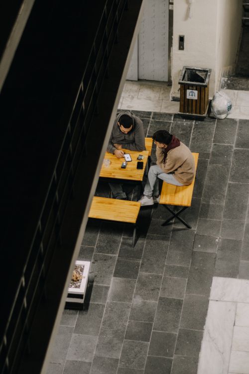 Free High-angle shot of two adults sitting at a wooden table on a paved urban floor, engaged in conversation. Stock Photo