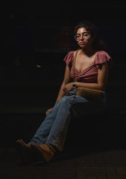 Fashionable young woman with eyeglasses sits on steps at night, showcasing casual attire.