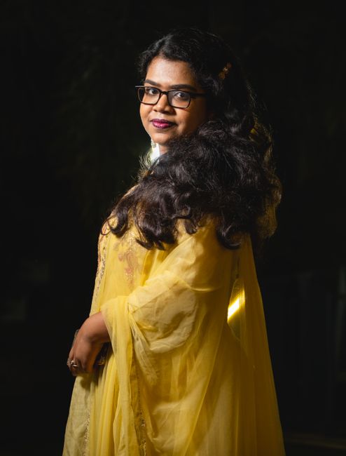 A woman in glasses and yellow traditional dress, posing gracefully.