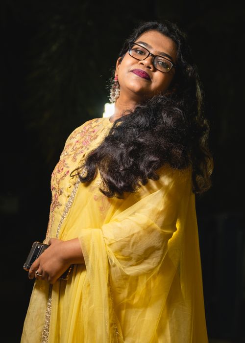 Free A young woman poses elegantly in a traditional yellow gown, adorned with embroidery and jewelry, smiling outdoors at night. Stock Photo