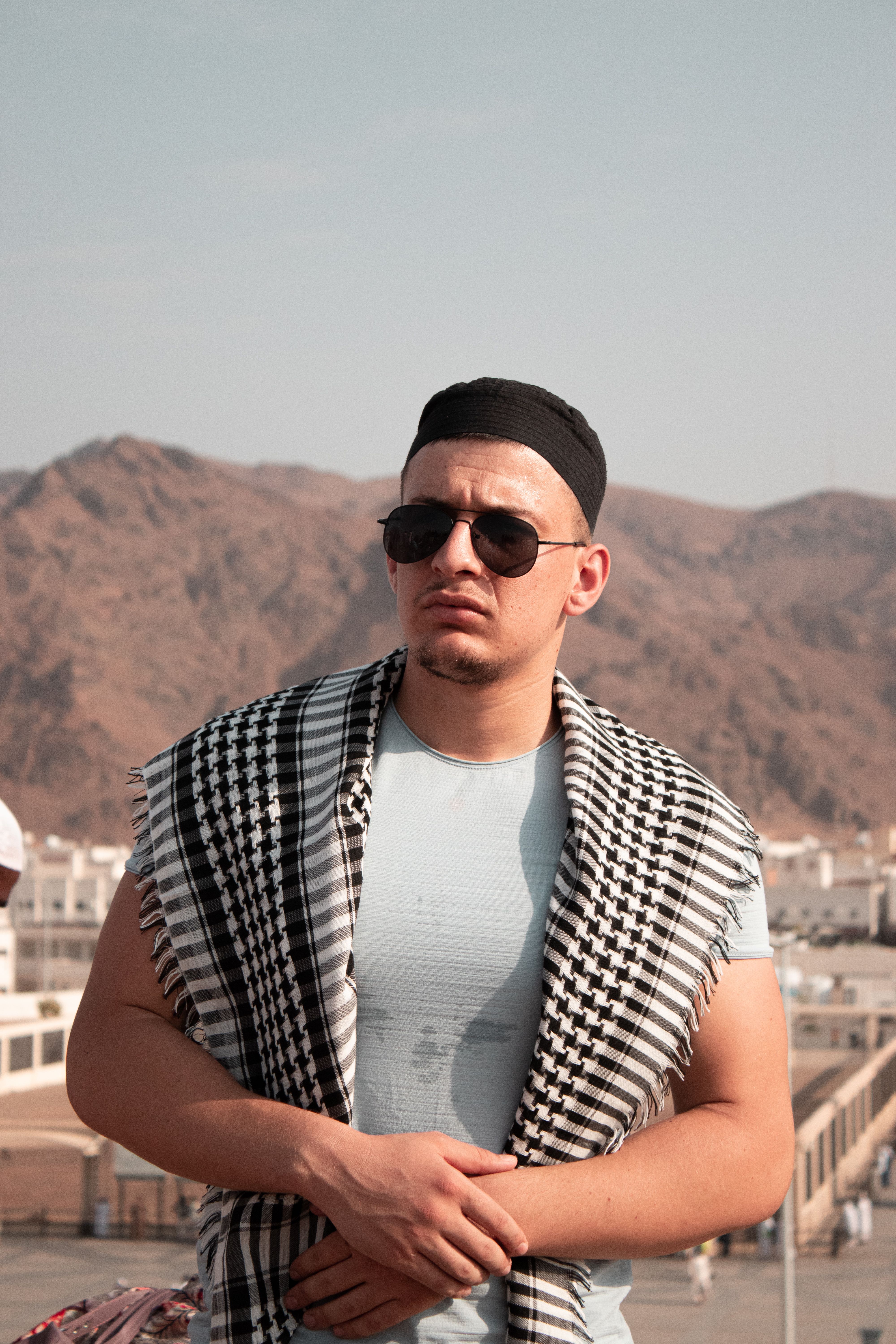 A confident man wearing sunglasses and a scarf poses outdoors against a mountain backdrop.