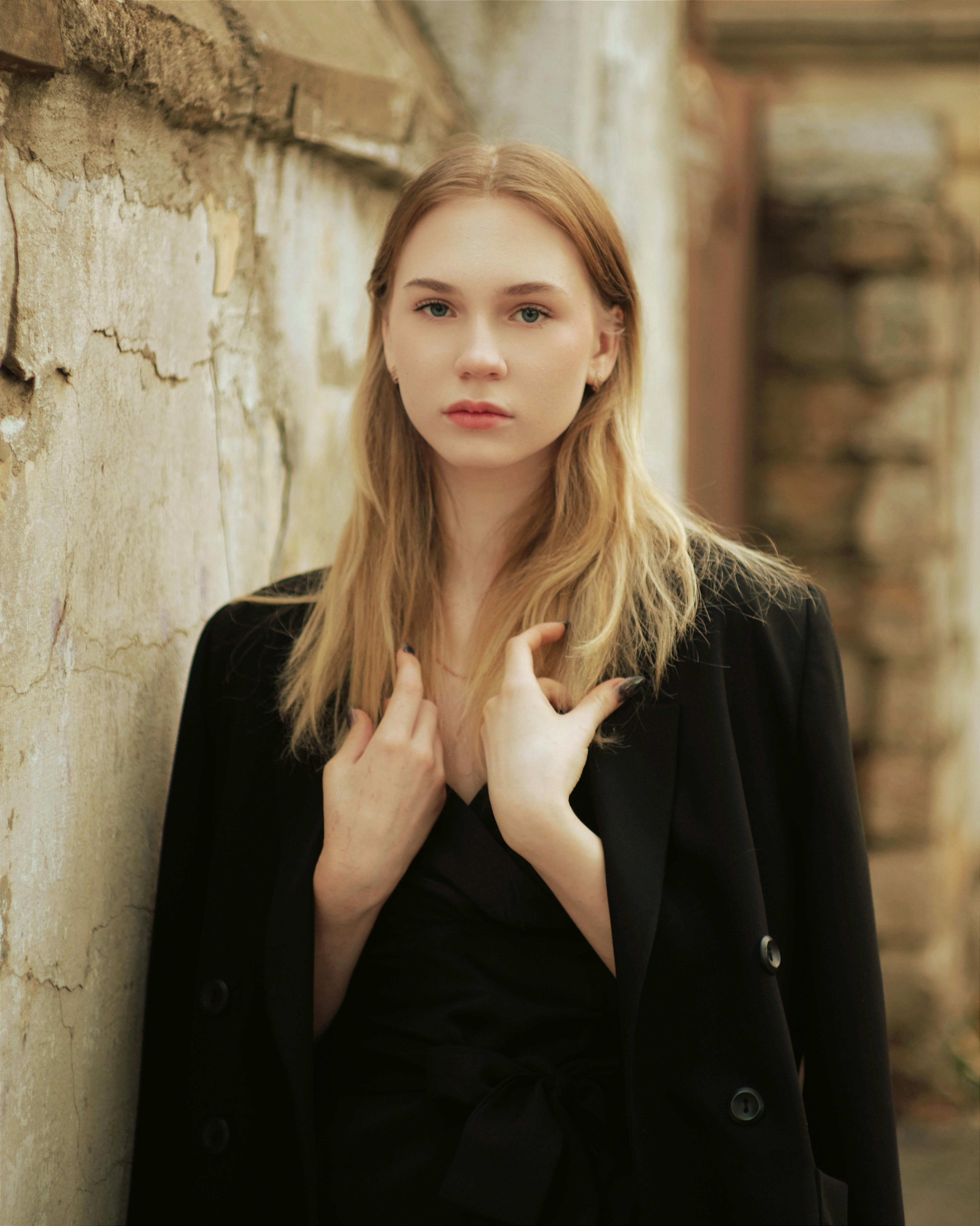 Blonde woman in black jacket posing against an old wall outdoors.