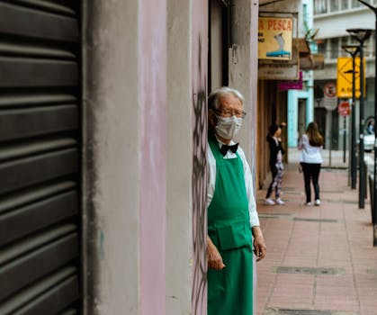 Elderly man in a green apron and mask stands by a wall on an urban sidewalk, observing passersby.