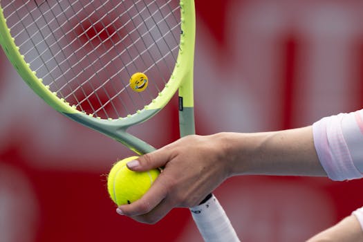 A hand holds a tennis ball by a racket with an emoji sticker, set against a sports backdrop.
