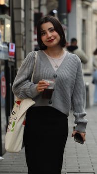 Smiling woman with a drink and tote bag, strolling down a lively urban sidewalk.