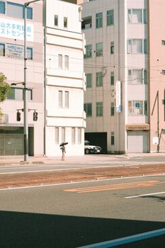 A woman with an umbrella walks along a sunlit city street near modern buildings.