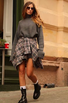 Fashionable woman in urban setting wearing sweater, boots, and sunglasses on city sidewalk.