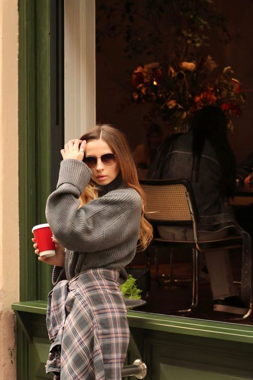 Free Stylish woman in a cozy sweater and sunglasses holding a coffee cup near a cafe window. Stock Photo