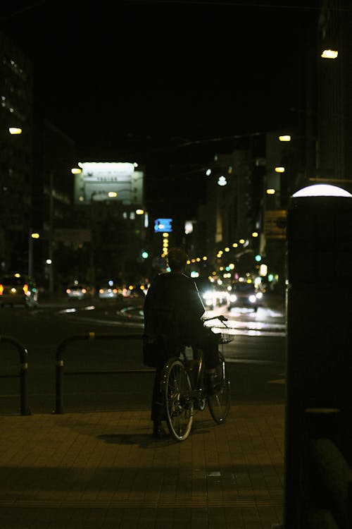 Person on a Bicycle Standing on the Sidewalk by the Street at Night