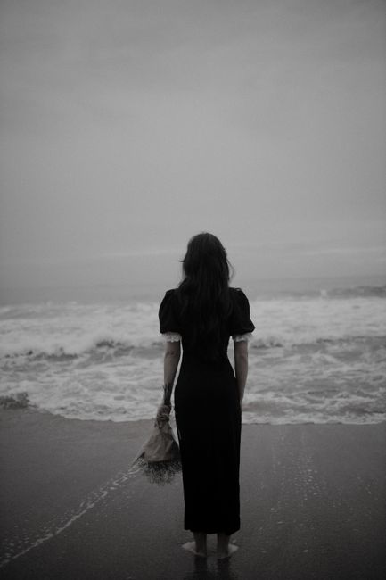 A woman in a black dress stands on the beach holding flowers, facing the sea.