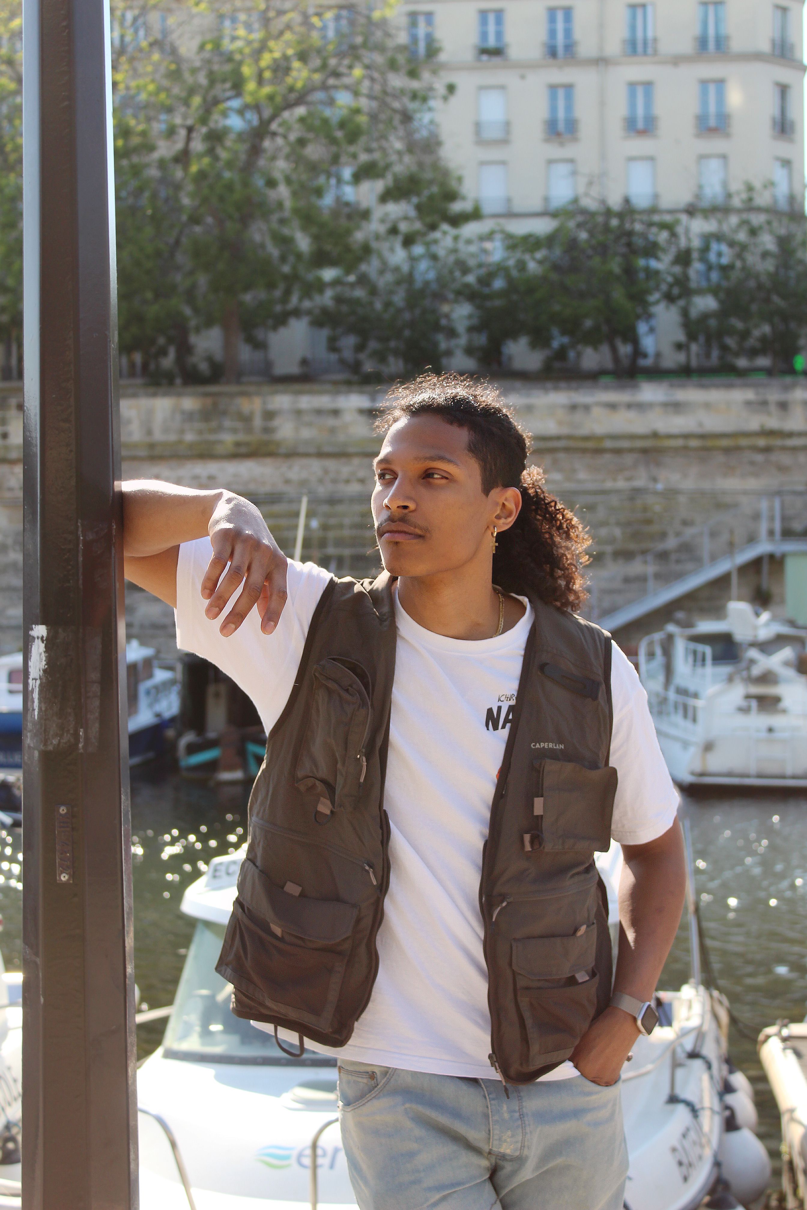 Man posing casually by urban canal, wearing vest and sunglasses.