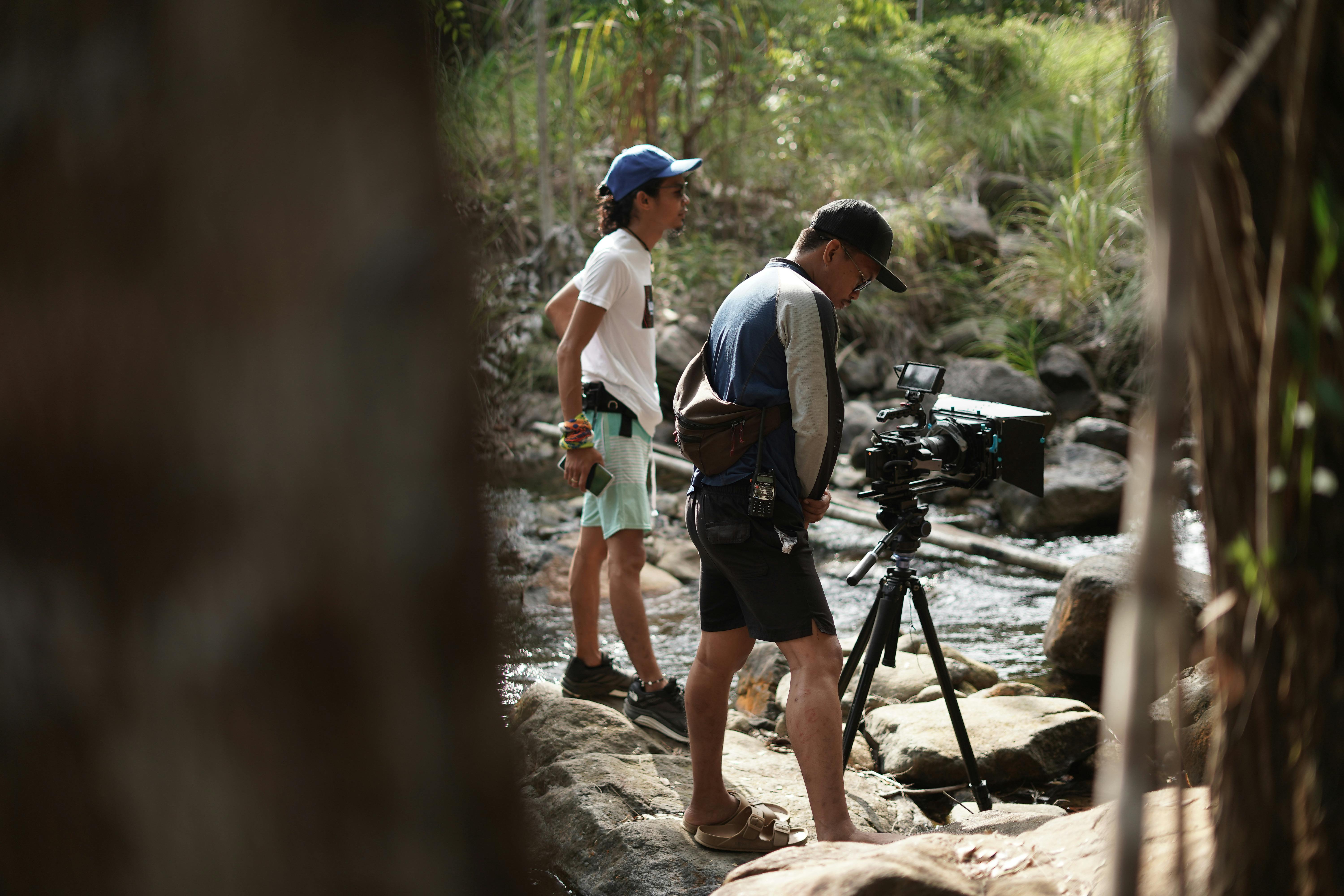 Two filmmakers prepare their camera equipment by a forest stream for a scene in natural surroundings.