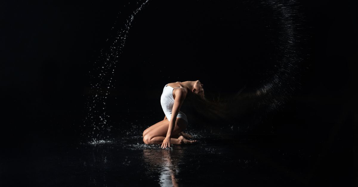 A woman gracefully dances with a water splash in a dramatic studio setting.