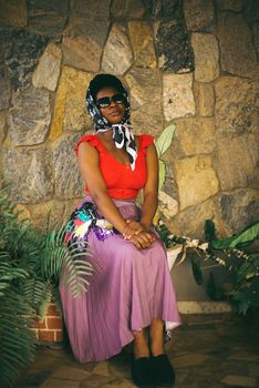 Stylish woman in vibrant attire posing seated against a stone wall with indoor plants.