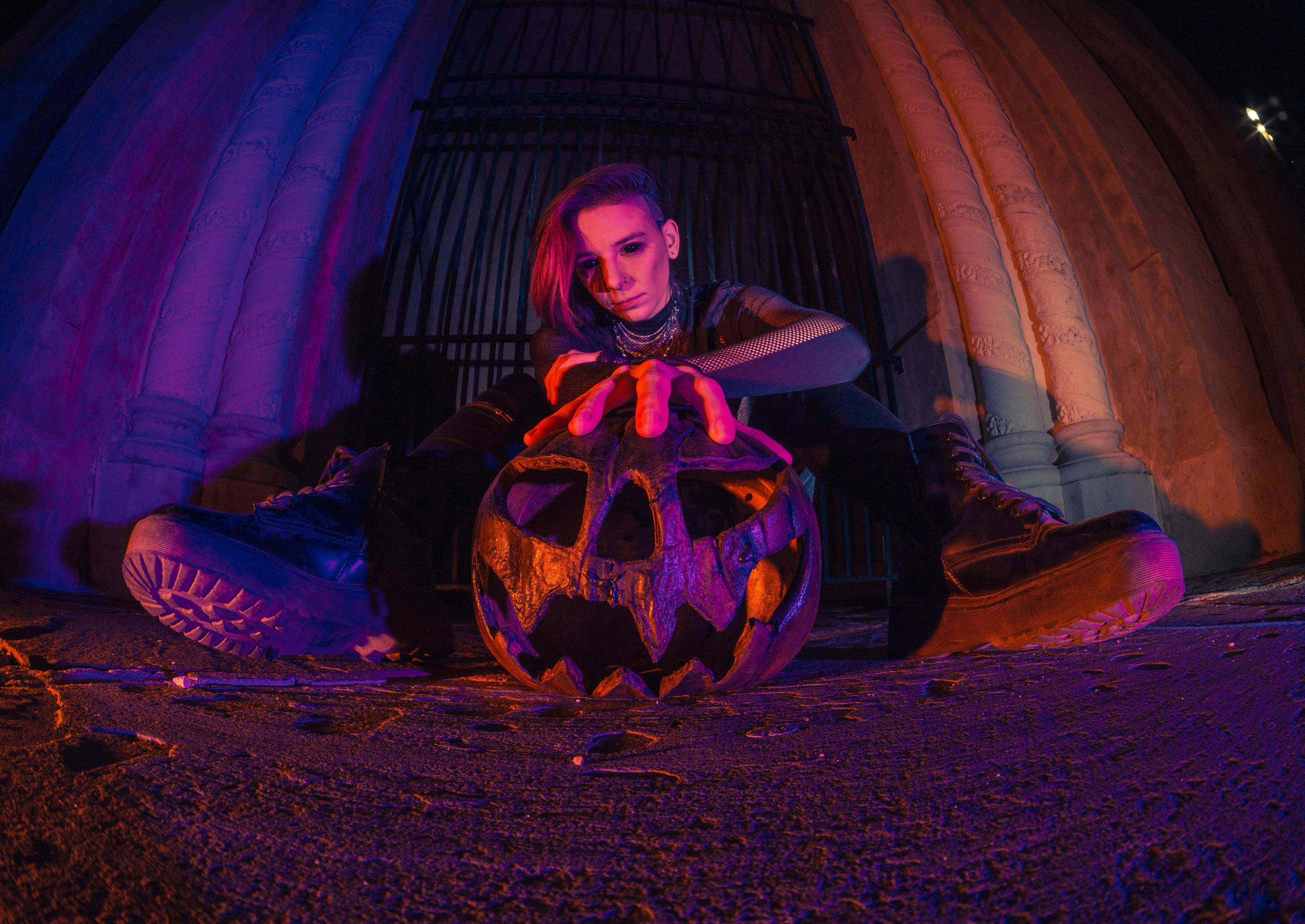 young woman in black leather boots and fishnet top posing with a dark jack o lantern