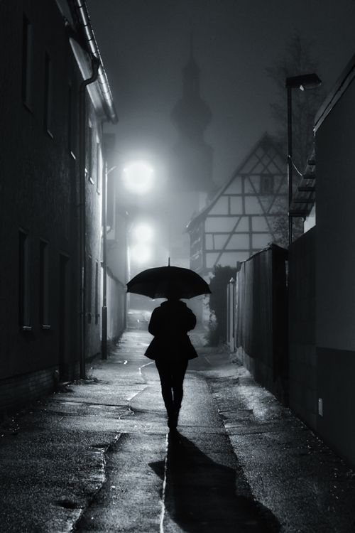 Free A solitary figure with an umbrella walking through a dimly lit, foggy alleyway at night. Stock Photo