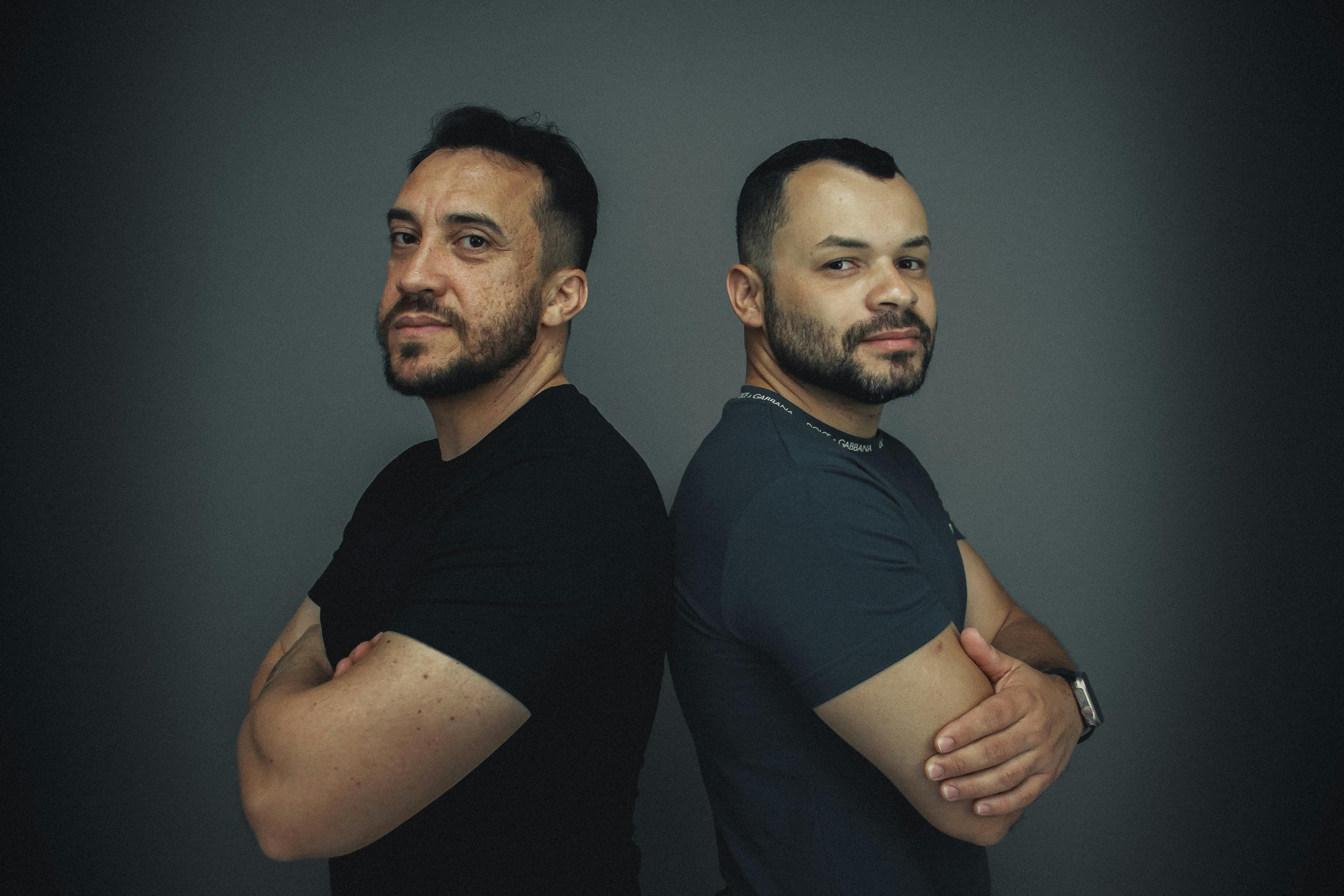 Fashion portrait of two confident men standing back to back against a gray studio background.