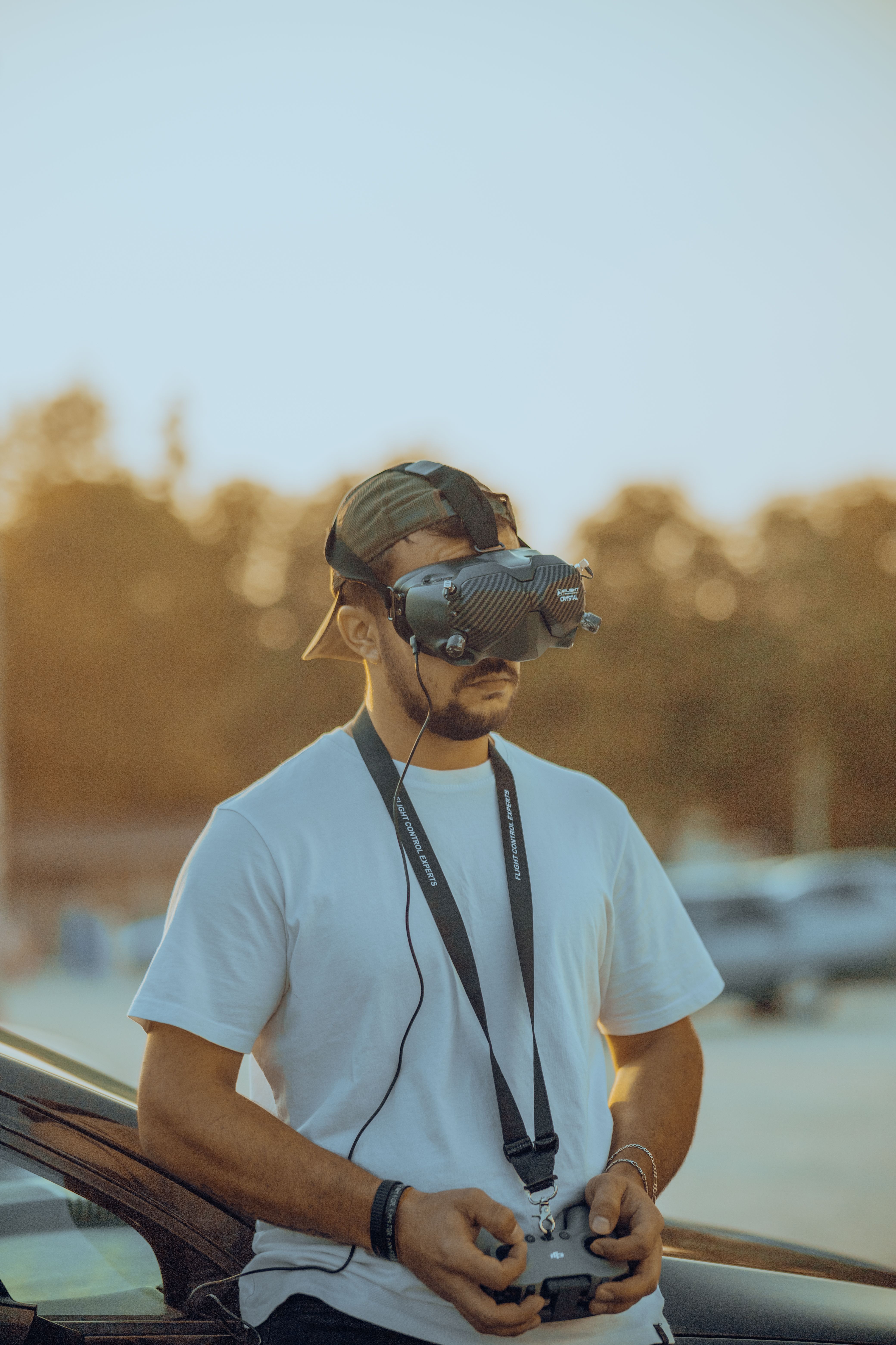 A man wearing VR headset and cap controlling a drone at a park during sunset.