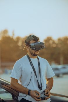 A man wearing VR headset and cap controlling a drone at a park during sunset.