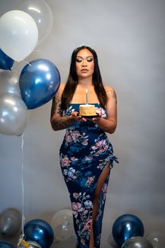 A woman in a floral dress holds a birthday cake with a candle among colorful balloons.