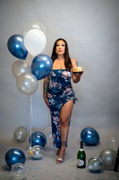 Woman in a floral dress holding a cake with balloons for a birthday celebration indoors.