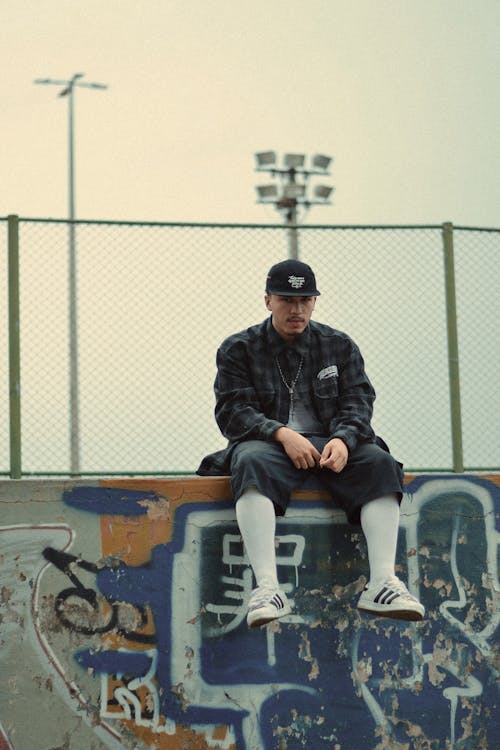 Man in Gray Flannel Shirt, Shorts, and Baseball Cap Sitting on a Wall