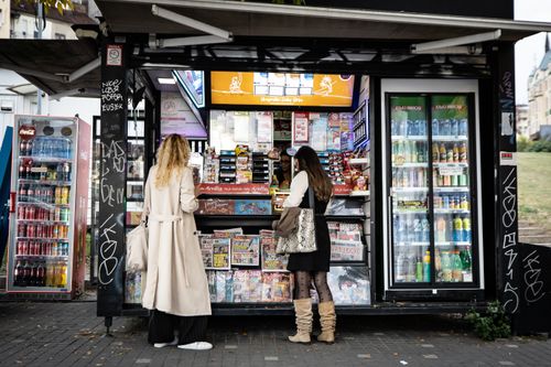 Free Women browsing at a street kiosk offering magazines and beverages in an urban setting. Stock Photo