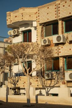 Urban apartment building facade with air conditioning units and barren trees in warm sunlight.