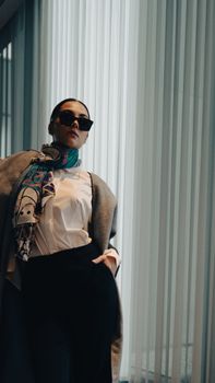 Elegant woman in sunglasses and scarf poses indoors with soft window lighting.