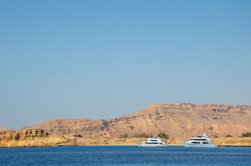 Free Luxurious yachts sail by a barren, desert coastline under a clear blue sky. Stock Photo