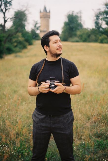 Man holding a vintage camera in a scenic Berlin field with a distant castle tower.