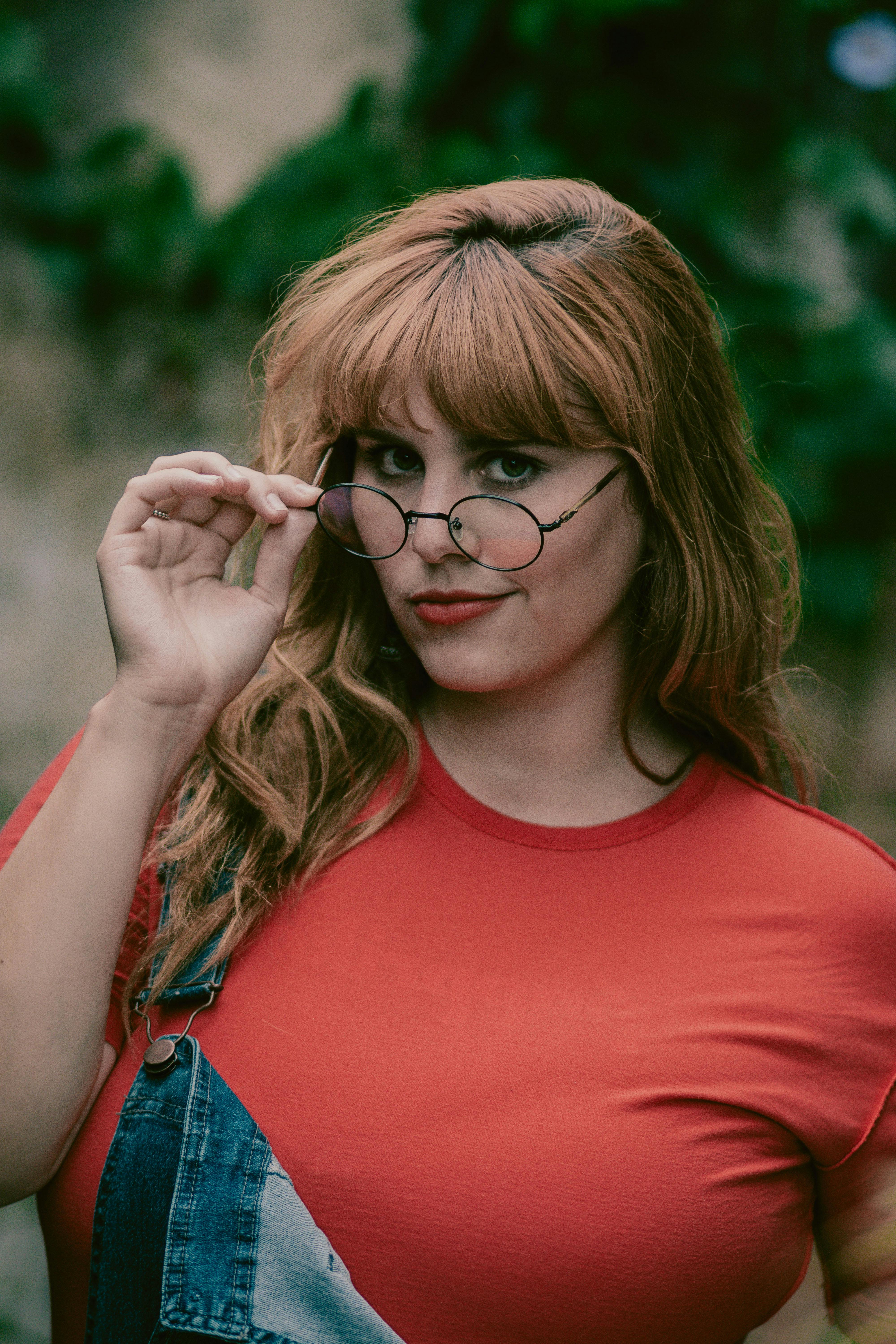 一位戴着圆眼镜、穿着红色衬衫的女人，在户外露出俏皮的表情微笑着