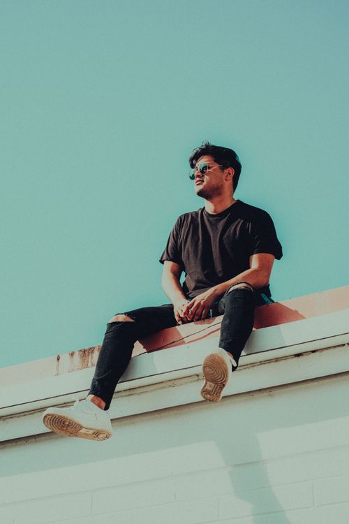 Free Trendy young man in sunglasses sitting on a rooftop against a bright sky in an urban setting. Stock Photo