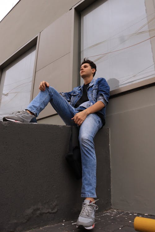 Young Man in a Denim Jacket and Jeans Sitting on a Wall