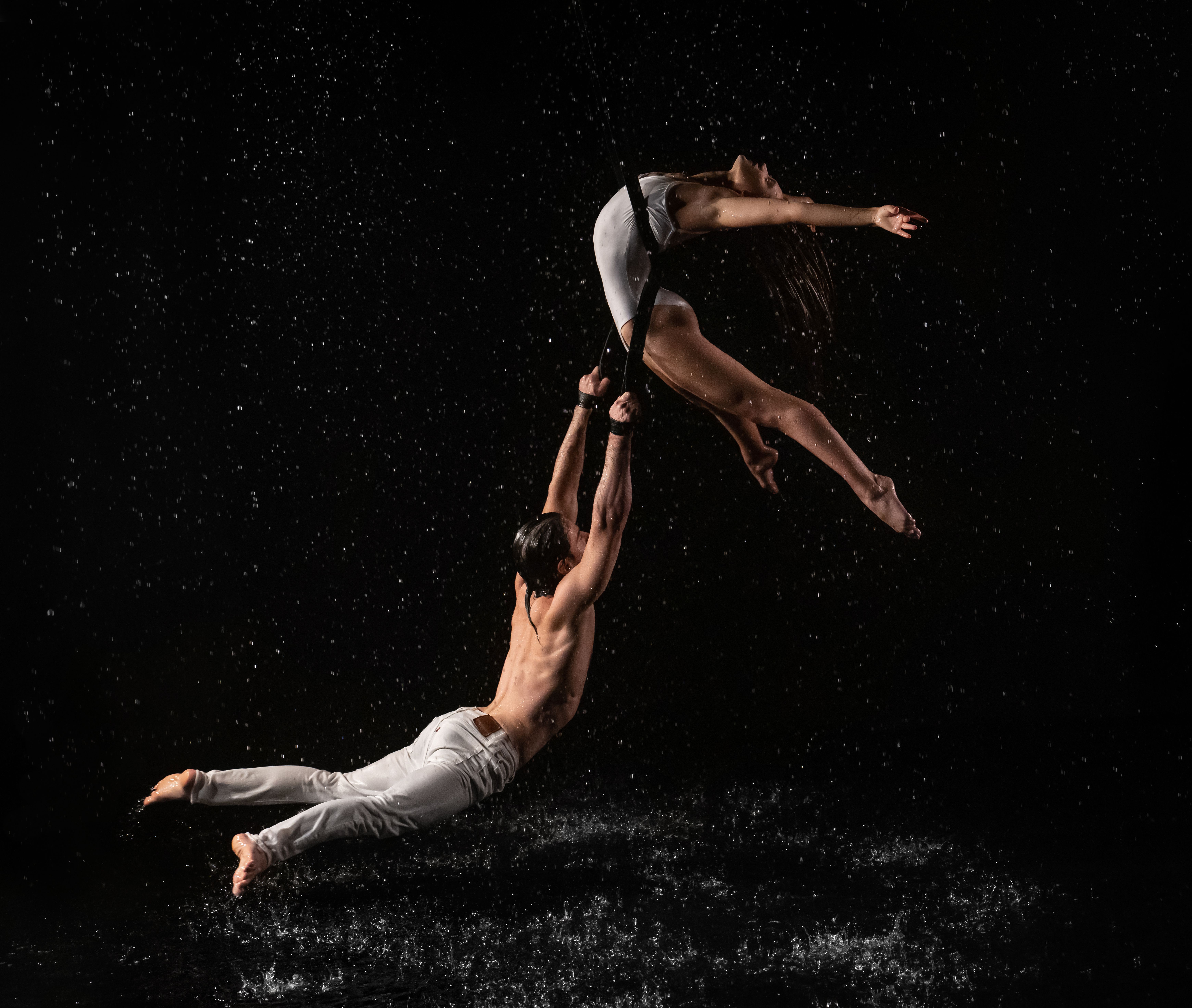 A man and woman perform aerial acrobatics on straps amidst water splash on black background.