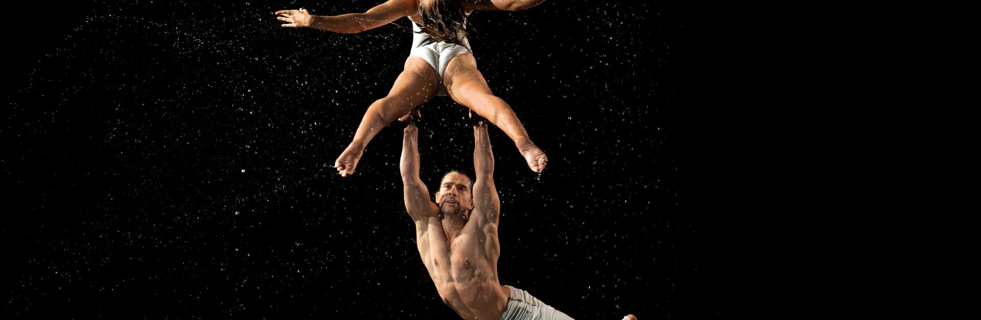 A mesmerizing duo performs aerial acrobatics amidst splashing water on a dark stage.
